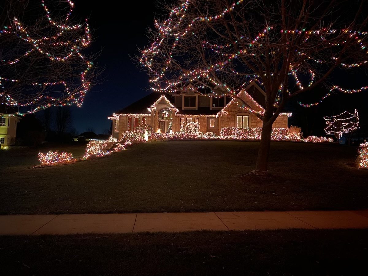 A stone ranch home at night with its rooflines lit and the big front-yard trees and shrubs wrapped in multicolor lights