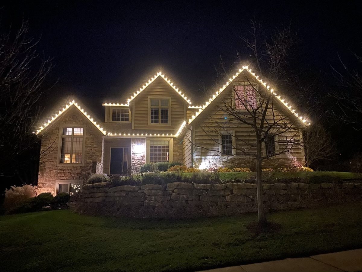 A stone and siding home with three steep gables traced in warm white C9 bulbs above a stone retaining wall