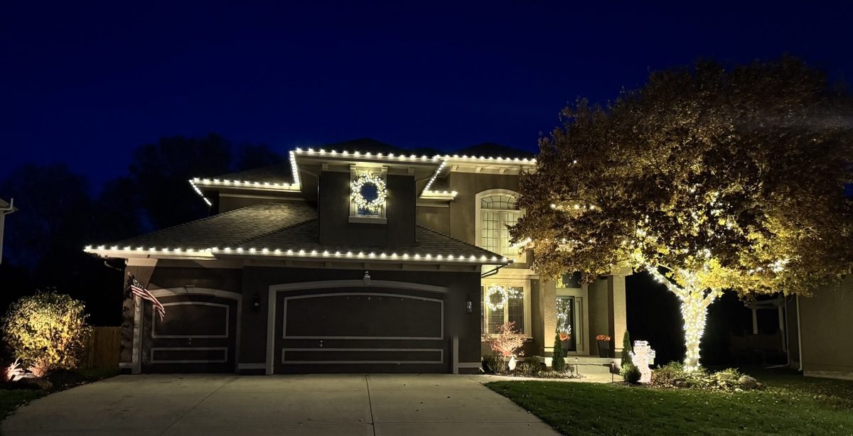 A two-story stucco home with warm white C9 bulbs along every roof edge, lit wreaths in the windows and a tree wrapped in lights
